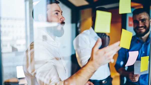 Smart smiling multiethnic business partners in elegant classical shirts speaking and looking at information on yellow stickers in window in office